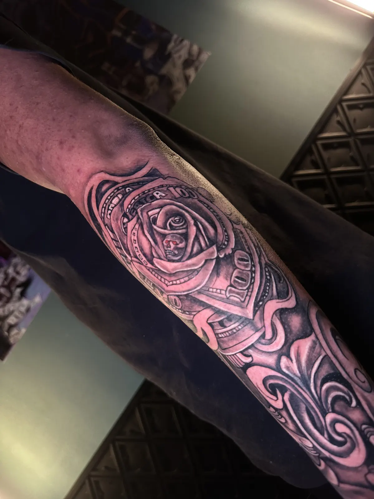 Tatuagem de rosa com detalhe de nota de cem reais no antebraço, peça com traço bem definido